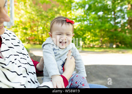 Laughing baby girl d'être tenue par la mère à l'extérieur à côté de feuilles vertes Banque D'Images