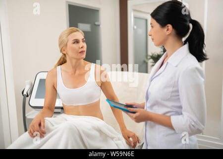 En attente de traitement. Femme sérieuse assis sur la table d'examen devant elle avant les procédures. cosmetologist Banque D'Images