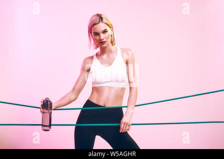 Fille de remise en forme. Jeune femme sportive en haut blanc et noir leggings holding bouteille d'eau et l'élaboration avec bande élastique debout contre fond rose en studio. Sport concept. L'équipement de sport Banque D'Images