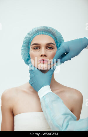 Traitement anti de vieillissement. Belle jeune femme en bleu medical hat en pointillés sur son visage d'avoir la chirurgie esthétique face à la chirurgie plastique. Plasticien en bleu gants holding scalpel. Concept de beauté. Banque D'Images