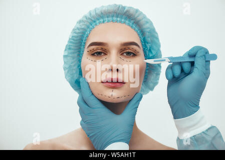 La chirurgie d'ascenseur de visage. Portrait de belle jeune femme en bleu medical hat cosmétique ayant du visage. Plasticien en bleu gants holding scalpel. Beauté concept Banque D'Images