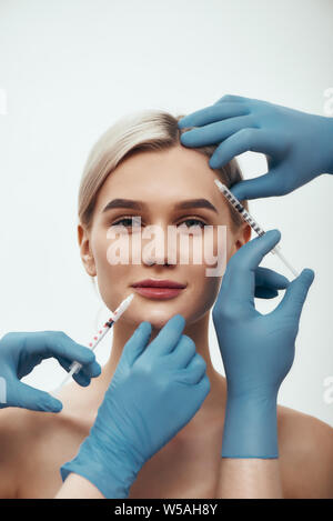 La création de la beauté. Portrait of young woman looking at camera et souriant alors que les médecins en bleu gants médicaux en injections dans son visage. Concept de Botox. La beauté. Chirurgie du visage