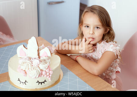 Magnifique petite fille debout dans la cuisine près de la table et des saveurs un gâteau d'anniversaire avec un doigt. Banque D'Images