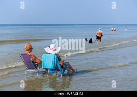 Deux dames âgées de soleil en vous détendre chaises / chaises de plage et de couple avec bassin pour chien dans l'eau le long de la côte de la mer du Nord pendant la vague de chaleur Banque D'Images