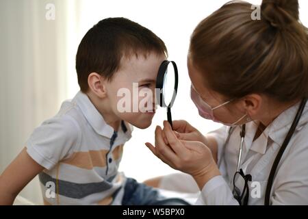 L'enfant fait une grimace et regarde le médecin à travers une loupe qu'une femme dans un manteau blanc tient dans ses mains. Banque D'Images