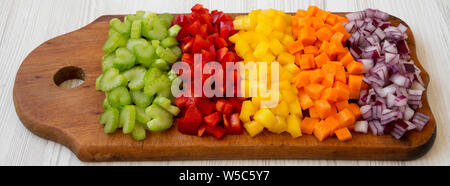 Les légumes frais, haché (carotte, céleri, oignon rouge, poivron) disposés sur une planche à découper sur un fond en bois blanc, vue de côté. Banque D'Images