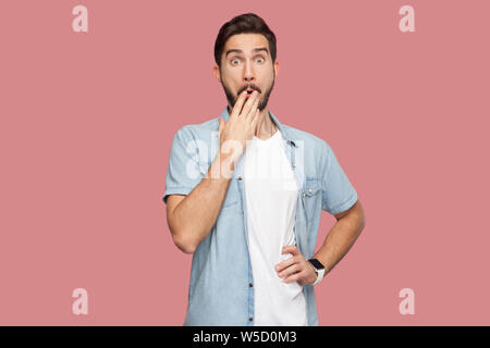 Portrait de jeune homme barbu choqué en bleu style casual shirt debout avec de grands yeux, couvrant sa bouche et à la recherche à l'appareil photo avec visage surpris. Banque D'Images