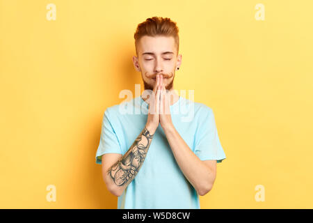 Homme barbu fidèle garde les mains et ferme les yeux, plus backgrouund isolé jaune. Jeune homme religieux en studio. de vie. conviction Banque D'Images