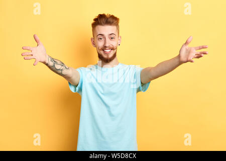 Young handsome smiling man wearing blue t-shirt debout avec les bras ouverts pour l'accolade sur fond jaune isolé. Bienvenue à un coiffeur. close up po Banque D'Images