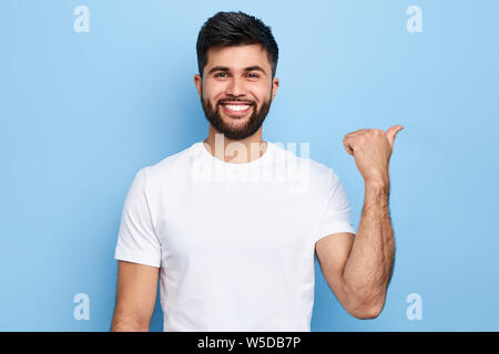 Guy Points de suite avec le pouce, être chaleureux et satisfait, indique la direction, porte des t-shirt blanc élégant, décontracté, isolé sur fond bleu.Pour Banque D'Images