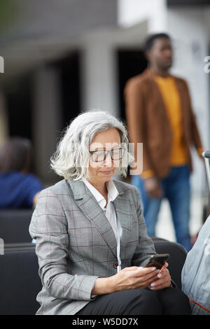 Senior business contenu dame avec les cheveux bouclés courts portant des lunettes et veste à carreaux texting on phone in airport Banque D'Images