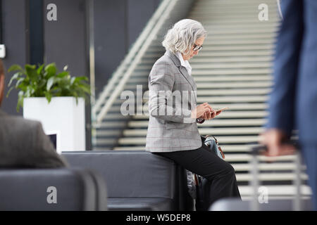 Les graves Asian businesswoman in formalwear assis à l'arrière du canapé dans le hall et le surf sur internet phone en attendant l'heure d'arrivée Banque D'Images