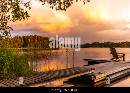 Des chaises longues en bois au coucher du soleil sur une jetée sur les rives du lac Saimaa calme en Finlande sous un ciel nordique avec un arc-en-ciel Banque D'Images