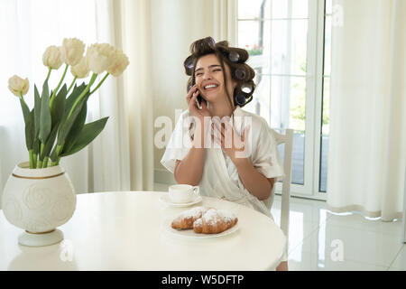 Belle jeune smiling brunette woman wearing sunglasses en parlant au smartphone pendant que boire du café et manger des croissants pour le petit déjeuner dans des Banque D'Images