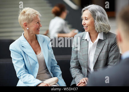 L'interracial mature positive mesdames dans jackets assis sur un canapé dans le hall et se parler gaiement Banque D'Images