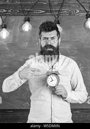 Enseignant dans les lunettes est titulaire d'un réveil. Régime de l'annexe et concept. Hipster barbu est titulaire d'réveil, tableau sur toile de fond, l'espace de copie. Homme avec barbe et moustache sur surpris face au réveil de pointage Banque D'Images