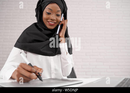 Belle, positive female office worker talking on phone, l'écriture, en souriant. Jeune africain femme musulmane portant le hijab en noir et blanc, travaillant en centre d'appel. Banque D'Images