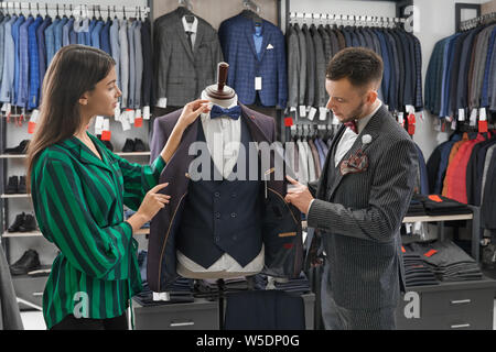 Shop aide cloosing expert-conseil complet pour les jeunes hommes d'affaires, à la recherche sur les vêtements au mannequin. Hôtel moderne et élégant vestes, chemises, gilets sur cintres et étagères dans la mode boutique. Banque D'Images