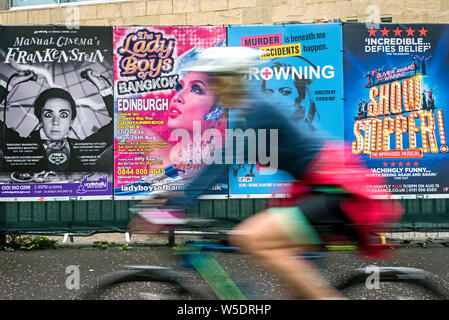 Un cycliste féminin rides par des affiches publicitaires pour Edinburgh Fringe Festival montre dans les prés, Édimbourg, Écosse, Royaume-Uni. (With motion blur) Banque D'Images