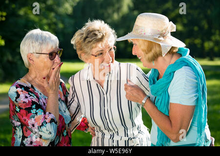 Trois femmes âgées commérage d'amis à l'extérieur, debout dans le parc. Choqué ou surpris femme âgée tenant sa main à sa bouche ouverte. Banque D'Images