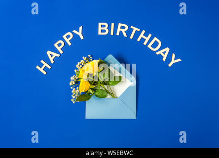 Roses jaune et blanc Gypsophila dans enveloppe bleue sur fond bleu. Joyeux anniversaire, texte des lettres en bois. Haut de la vue, télévision lay. Modèle pour Banque D'Images