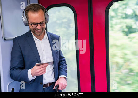 Businessman Standing On Train Film Streaming à partir d'un téléphone portable sur un casque sans fil Banque D'Images