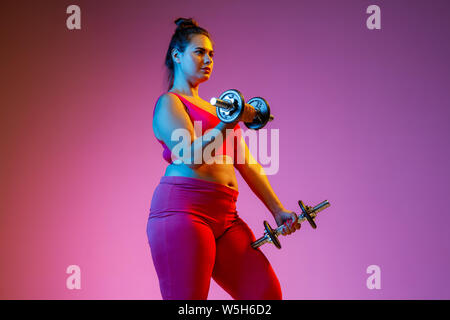 Young caucasian grande taille modèle féminin fait des exercices sur fond violet gradient de lumière néon. Sa formation le haut du corps avec des haltères. Concept du sport, mode de vie sain, positif. Banque D'Images