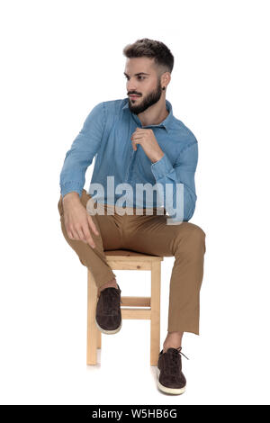 Dans l'homme détendu jeans bleu shirt assis avec le coude sur jambe tout en regardant ailleurs sur fond blanc Banque D'Images