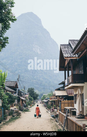 Muang Ngoi, Laos - Mai 2019 : femme laotienne en robe orange avec deux seaux de marcher sur la rue centrale du village Banque D'Images