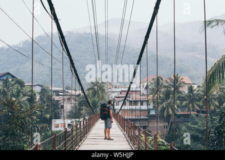 Sac à dos avec l'homme debout sur le pont suspendu en bois dans un village de montagne amoing jungles laotien au Laos. Banque D'Images