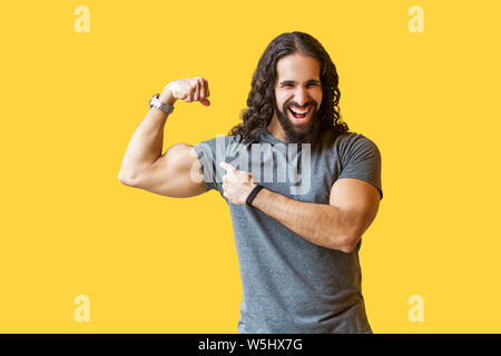 Portrait d'homme barbu fort drôle avec bodybuilder cheveux bouclés en gris tshirt debout, en montrant ses biceps et à pleines dents avec sourire. je Banque D'Images