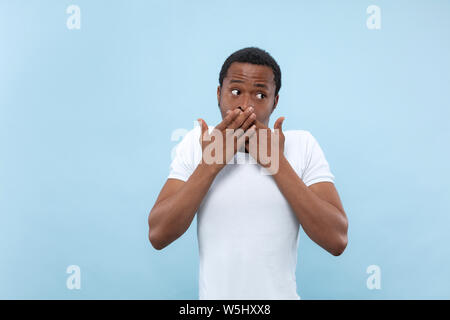 Demi-longueur close up portrait of young african-american man en chemise blanche sur fond bleu. Les émotions humaines, l'expression faciale, ad concept. Choqué, couvrant sa bouche de ses mains. Banque D'Images