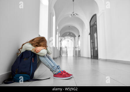 Vue latérale du triste school girl sitting on floor au corridor et se cacher la face en mains et pleurer. Fille blonde avoir mauvaise humeur terrible et marque après les cours. Concept de la dure période de l'adolescence. Banque D'Images