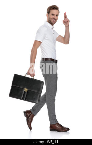 Side view of a smiling smart casual man holding briefcase et accueille avec un geste de la main à son front sur fond blanc Banque D'Images