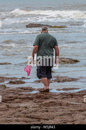 Grand homme d'âge moyen à la mer avec un filet de pêche rose dans sa main sur la plage. rockpooling Banque D'Images