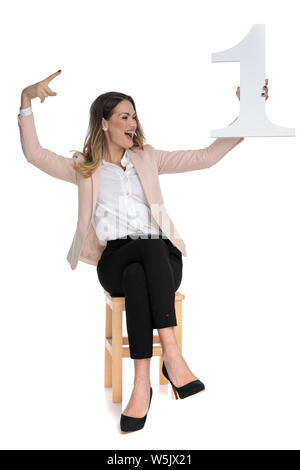 Happy businesswoman est titulaire d'un numéro et attend à l'autre tout en célébrant et assis sur fond blanc, pleine longueur photo Banque D'Images