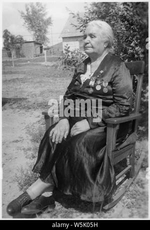 Une vieille femme indienne dans un fauteuil à bascule ; Portée et contenu : elle se trouve à l'extérieur, porte une robe avec quatre médailles épinglées sur le revers. Banque D'Images