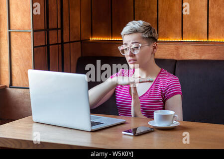 Time out ! Vue de côté portrait d'attention charmante fille indépendant avec des cheveux courts blonde est assise dans un café et faire appel vidéo sur ordinateur portable wit Banque D'Images