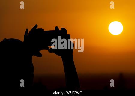 Silhouette de mains tenant un téléphone mobile et de faire de la photographie l'immense soleil pendant le coucher du soleil ou le lever du soleil. Homme fait une photo sur son téléphone Banque D'Images