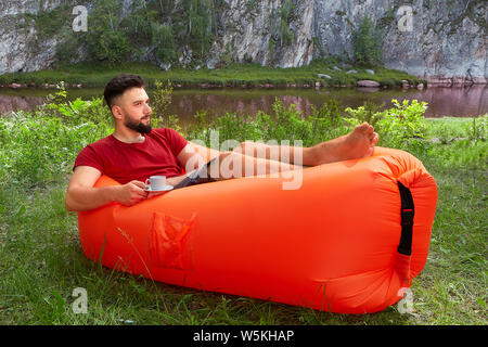 Young attractive caucasian man est allongé sur le canapé l'air orange avec une tasse de thé ou café dans sa main et se détendre pendant ses vacances. Banque D'Images