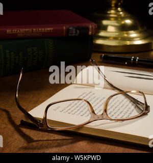 Une paire de lunettes portées à l'envers sur un livre ouvert, avec d'autres livres et d'un stylo d'encre sur un bureau avec une lampe en laiton. Banque D'Images