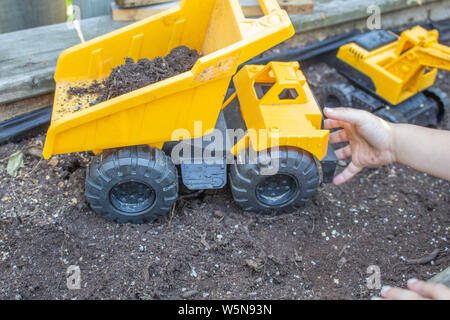 La main d'un jeune enfant atteint d'un jouet dump truck, assis sur le sol d'un parterre de fleurs. Banque D'Images