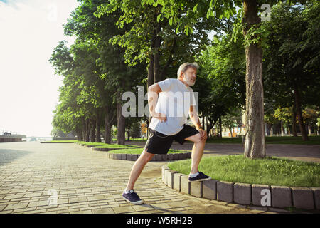 Man comme coureur à la rue de la ville. Modèle homme de race blanche et de jogging cardio training dans matin d'été. Faire des exercices d'étirement par près de prairie. Bonne hygiène de vie, sport, concept d'activité. Banque D'Images