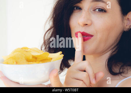 Pretty woman eating potato chips au lit à la maison avec le doigt sur ses lèvres Banque D'Images