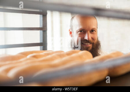 Portrait de jeune adulte baker avec longue barbe en uniforme blanc debout dans son milieu de travail et à la recherche sur le pain frais dans le bac, sourire à pleines dents, Indo Banque D'Images