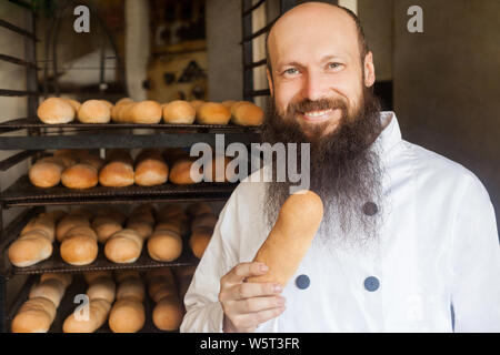 Portrait de jeunes adultes satisfaits chef avec longue barbe en uniforme blanc debout dans sa boulangerie et la tenue du pain frais, looking at camera avec dents Banque D'Images