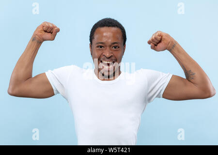 Demi-longueur close up portrait of young african-american man en chemise blanche sur fond bleu. Les émotions humaines, l'expression faciale, ad, concept. La célébration, se demande, étonné, choqué, fou heureux. Banque D'Images