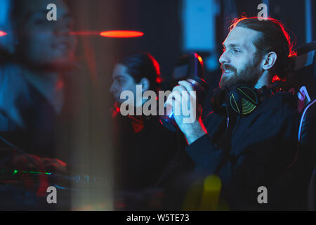 Sourire satisfait les jeunes gamer barbu avec un casque sur le cou assis dans un fauteuil confortable et de boire des boissons énergétiques tout en jouant le jeu vidéo Banque D'Images