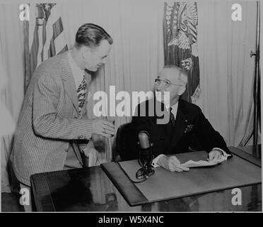 Le président Harry S. Truman assis à un bureau, souriant à un assistant, au moment de son annonce de la fin de la Seconde Guerre mondiale en Europe. Banque D'Images