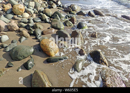 Différentes formes et tailles de roches, pierres et cailloux en couleurs de corail, de jade, de rouille, et tan en plein soleil éparpillés sur une plage de sable brun foncé Banque D'Images